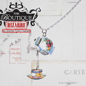 May include: A silver chain necklace with a teapot and teacup charm. The teapot and teacup are decorated with floral patterns in shades of blue, pink, and yellow. The necklace is labeled "The Boutique Bizarre" and "By: Kristin Torres Gurule".