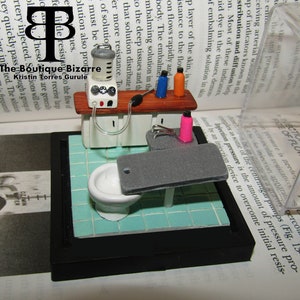 May include: A miniature model of a medical embalming room with a white toilet, a gray table, and a small cabinet with a pump and bottles. The room is set on a blue and white tiled floor.