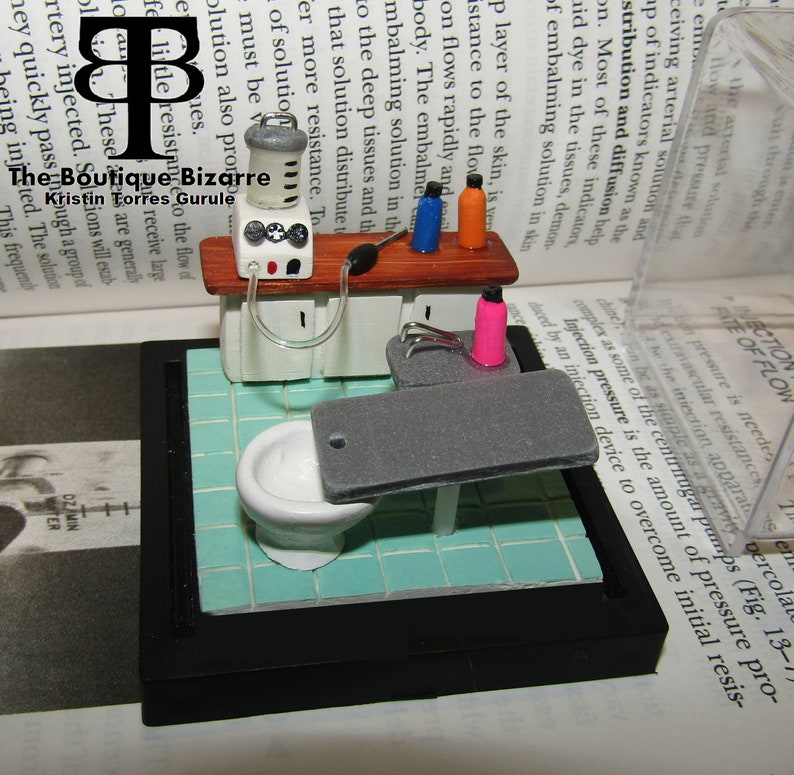 May include: A miniature model of a medical embalming room with a white toilet, a gray table, and a small cabinet with a pump and bottles. The room is set on a blue and white tiled floor.