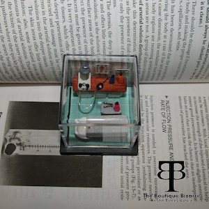 May include: A miniature diorama of a medical embalming room. The diorama is encased in a clear plastic cube with a teal tiled floor. The room features a small table with a miniature embalming machine, a sink, and a pair of pliers. The diorama is a unique and detailed representation of a medical procedure.