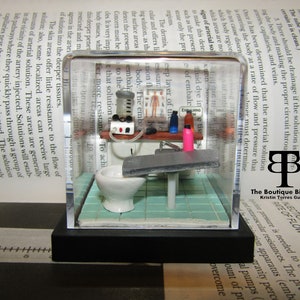 May include: A miniature diorama of an embalming room. The diorama is enclosed in a clear acrylic cube with a black base. The diorama features a small white toilet, a sink, a table with a bottle of pink fluid, and a sign that reads "Embalming Room".