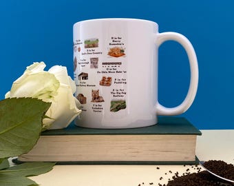May include: White ceramic mug featuring a colorful design with the alphabet and various images. The mug is placed on a closed book with a white rose and green leaves. The background is a solid blue.