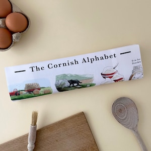 May include: A white tea towel with a black and white illustration of a hay wagon, a black bear, and a bowl of cream. The text "The Cornish Alphabet" is printed in black at the top of the towel. The text "D is for Dreckly" is printed in black in the upper right corner of the towel.