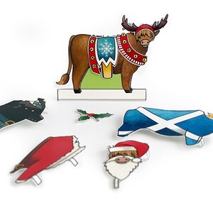May include: A brown and white reindeer with antlers, wearing a red and gold Christmas-themed harness with bells. The reindeer is standing on a green and white platform. There are also paper cutouts of a Santa Claus head, a holly sprig, a kilt, a red hat, and a Scottish flag.