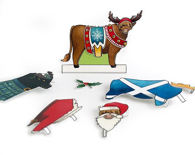 May include: A brown and white reindeer with antlers, wearing a red and gold Christmas-themed harness with bells. The reindeer is standing on a green and white platform. There are also paper cutouts of a Santa Claus head, a holly sprig, a kilt, a red hat, and a Scottish flag.