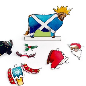May include: A set of paper craft cutouts featuring a Scottish Highland cow, a Father Christmas, a reindeer, a Christmas tree, and a Scottish kilt. The cutouts are printed on white paper with colourful details.
