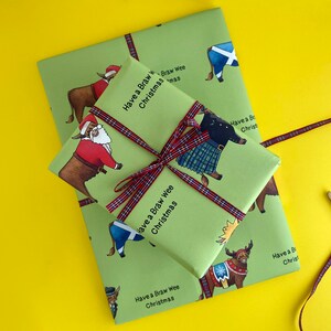 May include: Green wrapping paper with a Scottish Highland cow design. The paper features the text "Have a Braw Wee Christmas" repeated throughout. The paper is tied with a red and black tartan ribbon.