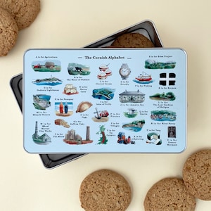 May include: A rectangular tin with a light blue lid featuring illustrations of the Cornish Alphabet. The tin is open, revealing cookies inside and around it. The tin's design includes various Cornish landmarks and symbols.