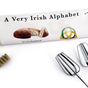 以下が含まれることがあります： 「A Very Irish Alphabet」という文字と、伝統的なアイルランドのフルーツケーキであるバーンブラックのイラストが描かれた白いキッチンタオル。イラストには「B is for Barmbrack」という文字も書かれている。キッチンタオルには、緑、黄色、白のケルトの結び目も描かれている。