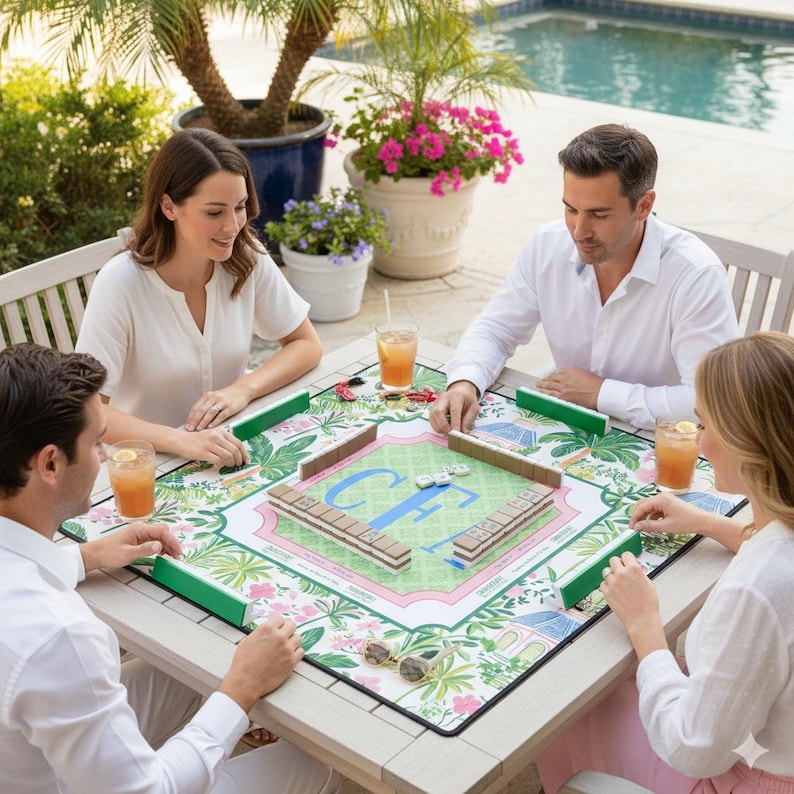 A group of four people playing mahjong outdoors. The game is set on a white table with a floral patterned mat. The mat features green, pink, and white colors. The players are enjoying drinks.