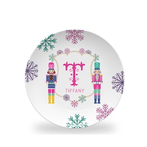 May include: A white ceramic plate with a pink, gold, and blue design featuring two nutcracker figures, snowflakes, and the letter "T" with the name "TIFFANY" below.