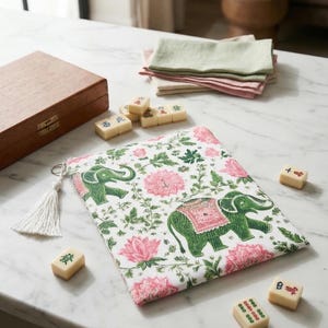 Op de afbeelding: Een witte tas met een roze bloemen- en groene olifantenpatroon, een witte kwast en verschillende mahjongtegels liggen op een marmeren oppervlak. Een houten kist en gevouwen servetten zijn ook zichtbaar.