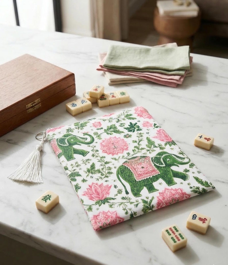 Op de afbeelding: Een witte tas met een roze bloemen- en groene olifantenpatroon, een witte kwast en verschillende mahjongtegels liggen op een marmeren oppervlak. Een houten kist en gevouwen servetten zijn ook zichtbaar.
