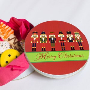 May include: A round red tin with a green ribbon and the words "Merry Christmas" printed on it. The tin features a row of six nutcracker figurines in various colors and designs.