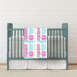 May include: White baby blanket with a repeating pattern of the name "Madison" in pink, blue, and green lettering. The blanket is displayed on a crib with a white sheet.