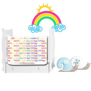 May include: A white crib with a rainbow and clouds printed on the headboard. The crib has a white mattress with a colorful print that says "After every storm there is a rainbow of... Angel Beautiful Strong Sweet Joyful Blessing Angel Beautiful" and "I am! Alexandra".