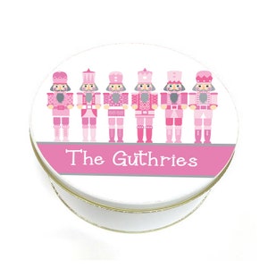 May include: A white round tin with a pink and gray border. The tin features a row of six pink nutcracker figurines with gray faces and gold crowns. The text "The Guthries" is printed in gray on the pink border.
