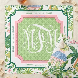 Op de afbeelding: Een vierkant spelbord in groen en roze met een wit monogram en bloemenrand, naast een bijpassend etui. Het bord heeft tekst langs de randen en het etui toont een tropische sc&egrave;ne. Er liggen verschillende speltegels verspreid.