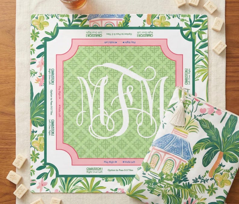 Op de afbeelding: Een vierkant spelbord in groen en roze met een wit monogram en bloemenrand, naast een bijpassend etui. Het bord heeft tekst langs de randen en het etui toont een tropische sc&egrave;ne. Er liggen verschillende speltegels verspreid.