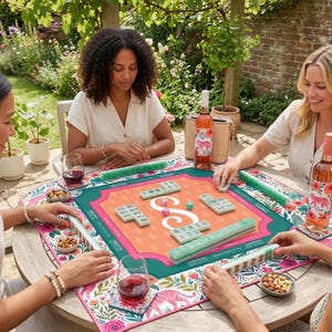 May include: A group of people playing mahjong outdoors. The game is set on a floral patterned mat with a pink and orange center. Wine bottles and glasses are on the table. The game pieces are green and white.