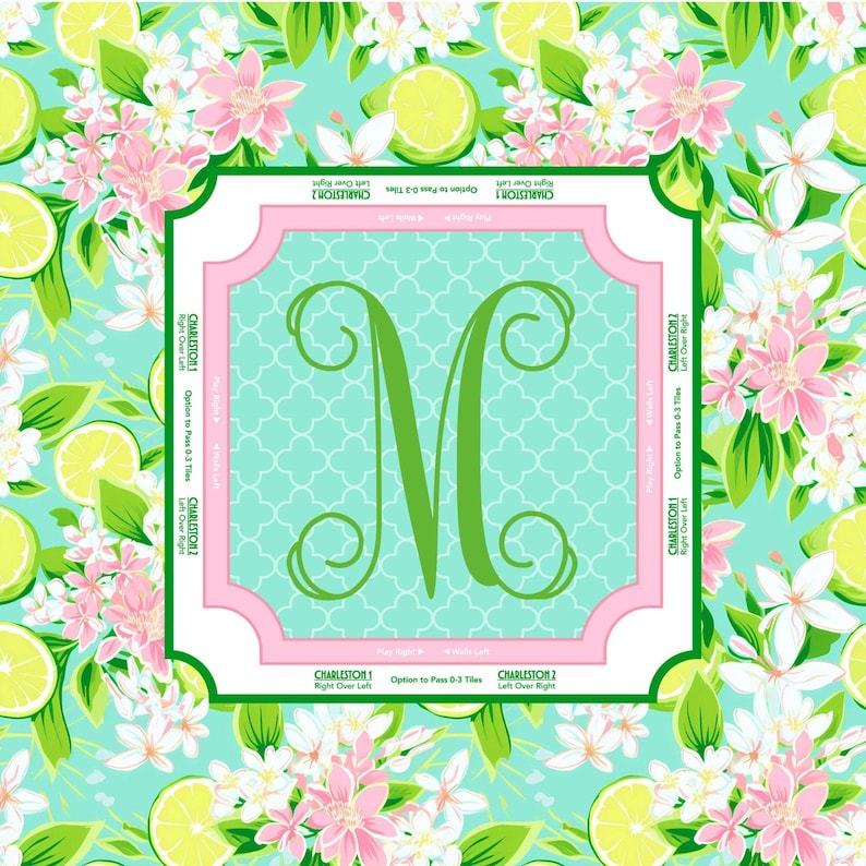 May include: A square, decorative item featuring a large green letter "M" on a light blue background with a white trellis pattern. The item is framed in pink and surrounded by a floral pattern of pink flowers, green leaves, and lime slices.
