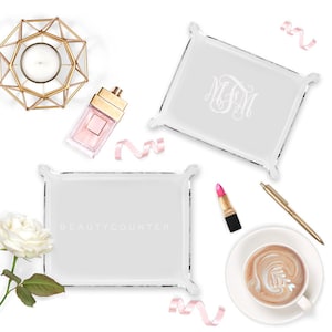 May include: Two clear acrylic trays with raised edges. The top tray has the monogram "JFM" etched in white. The bottom tray has the text "BEAUTYCOUNTER" printed in black. Both trays are on a white background with decorative items around them.