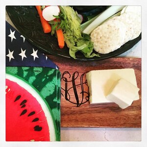 May include: A wooden cutting board with a monogrammed design and a block of white cheese. A plate with celery sticks, carrots, radishes, and crackers is in the background.