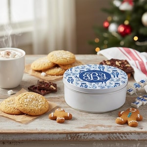 May include: A white cookie tin with a blue and white floral design and the letter "B" on top. The image also shows cookies, a cup of hot chocolate, and a Christmas tree in the background.