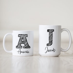 May include: Two white ceramic mugs with black sketched letters and names. The mug on the left has the letter "A" and the name "Amanda". The mug on the right has the letter "J" and the name "Jacob".