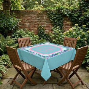 May include: A square, light blue tablecloth with a pink and blue floral design and a pink central pattern, set on a wooden table. The table is surrounded by wooden chairs and set in a garden setting.