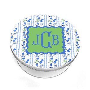 May include: A round, white metal tin with a decorative lid. The lid features a green scalloped rectangle with the blue letters "JCB". The background has blue and white stripes and small blue floral designs.