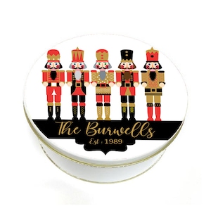 May include: Round white tin with a black and gold design featuring five nutcracker figures. The text "The Burwells Est. 1989" is printed on the tin.