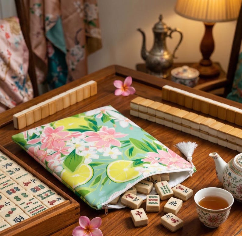 Op de afbeelding: Een mahjongset met houten tegels en een etui met bloemenprint. De etui is lichtblauw met roze bloemen en gele citroenen. De set bevat een houten kist, tegels en een kopje thee.