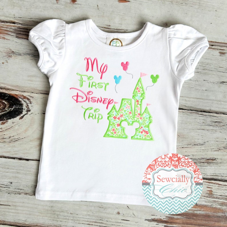 May include: White short-sleeved t-shirt with the embroidered text "My First Disney Trip" in pink and green. The design includes a castle with a Mickey Mouse silhouette and balloons.