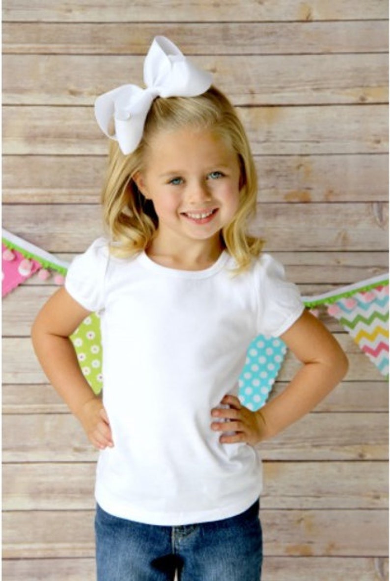 May include: A young girl wearing a white t-shirt with a large white bow in her hair. She is standing in front of a wooden background with colorful flags.