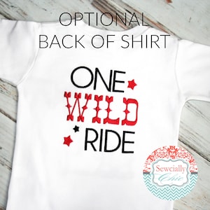 K&ouml;nnte beinhalten: Wei&szlig;es T-Shirt mit schwarzer und roter Stickerei, auf dem "ONE WILD RIDE" mit roten Sternen steht. Das Shirt liegt auf einem wei&szlig;en Holzgrund.