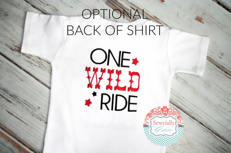 K&ouml;nnte beinhalten: Wei&szlig;es T-Shirt mit schwarzer und roter Stickerei, auf dem "ONE WILD RIDE" mit roten Sternen steht. Das Shirt liegt auf einem wei&szlig;en Holzgrund.