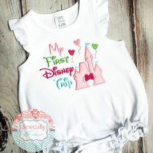 May include: White baby romper with ruffled sleeves and hem. The romper features the embroidered text "My First Disney Trip" in pink, green, and blue, along with a pink castle and heart-shaped balloons. The size is 6-12M.