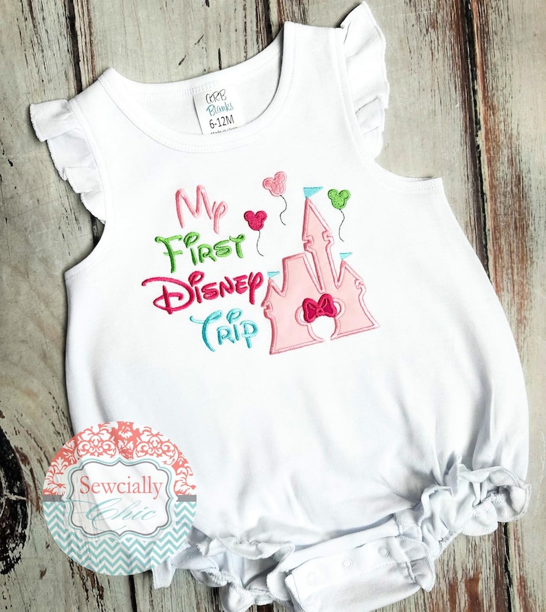 May include: White baby romper with ruffled sleeves and hem. The romper features the embroidered text "My First Disney Trip" in pink, green, and blue, along with a pink castle and heart-shaped balloons. The size is 6-12M.