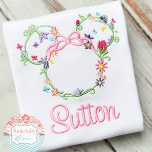 May include: White t-shirt with a pink floral embroidered design in the shape of Mickey Mouse ears. The name "Sutton" is embroidered in pink below the design.