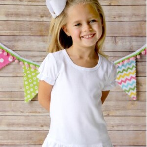 May include: A white short-sleeved t-shirt with a ruffled hem. The shirt is made of a soft, comfortable fabric and has a round neckline. The girl is wearing blue jeans and a white bow in her hair.