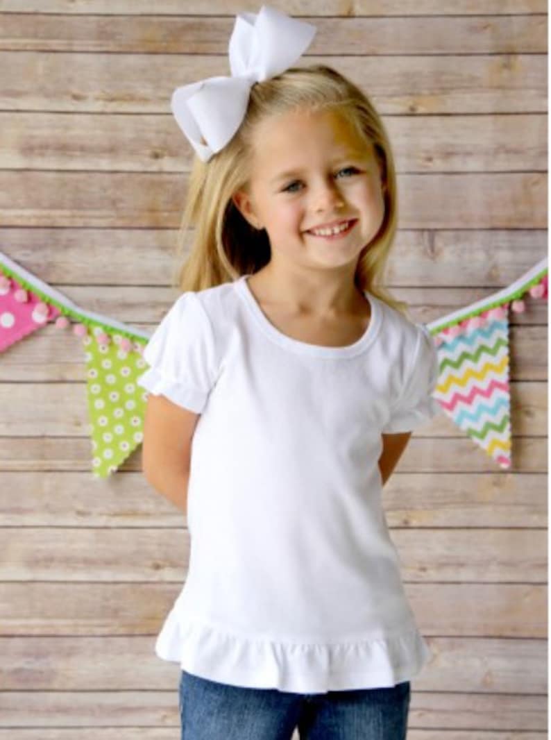 May include: A white short-sleeved t-shirt with a ruffled hem. The shirt is made of a soft, comfortable fabric and has a round neckline. The girl is wearing blue jeans and a white bow in her hair.