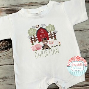 May include: White baby onesie with an embroidered farm scene featuring a red barn, a horse, a pig, and a sheep. The onesie has the name "CHRISTIAN" embroidered below the farm scene.