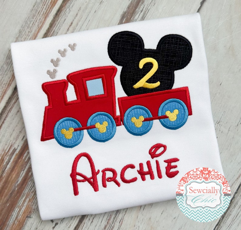 May include: Red and blue embroidered train with a black Mickey Mouse head on top. The train has a yellow number 2 on the Mickey Mouse head. The name Archie is embroidered in red below the train.