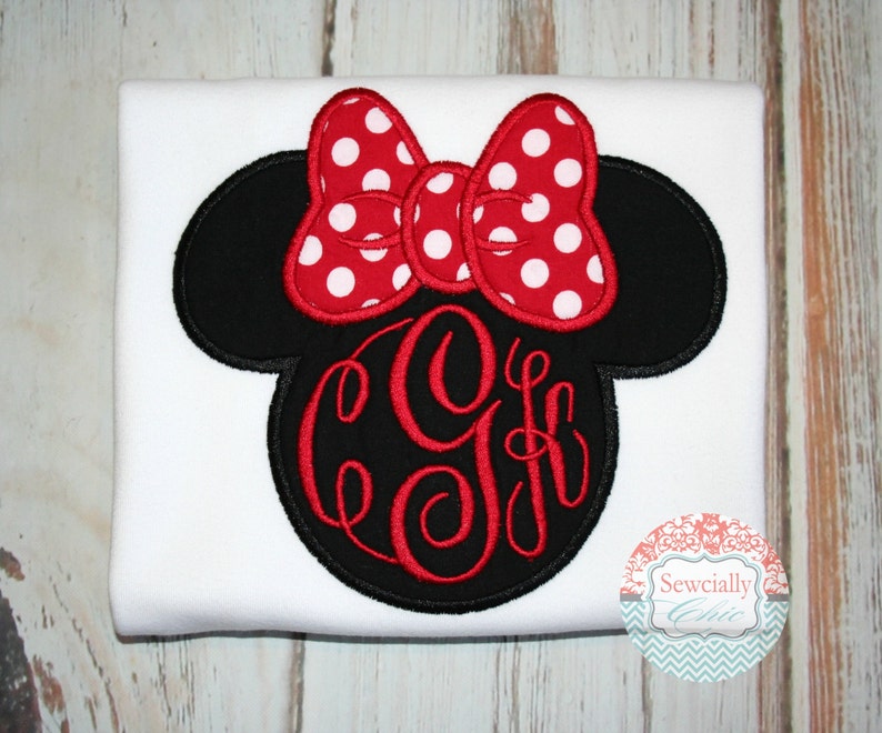 May include: White t-shirt featuring a black and red embroidered Minnie Mouse head with a red polka dot bow and a red monogram.