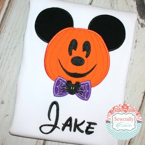 Puede incluir: Una camiseta blanca con una calabaza naranja bordada, orejas y rasgos faciales negros de Mickey Mouse. Debajo de la calabaza hay una pajarita morada con un patr&oacute;n de telara&ntilde;a y una peque&ntilde;a ara&ntilde;a negra. El nombre Jake est&aacute; bordado en color negro.