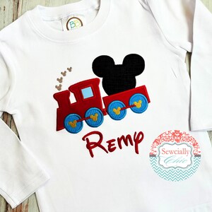 May include: White long-sleeved shirt with an embroidered red train with blue wheels and a black Mickey Mouse head. The train has gold Mickey Mouse ears on the wheels. The shirt has the name "Remy" embroidered in red below the train. The shirt also has a small circular label with the text "Sewcially Chic" in a decorative font.