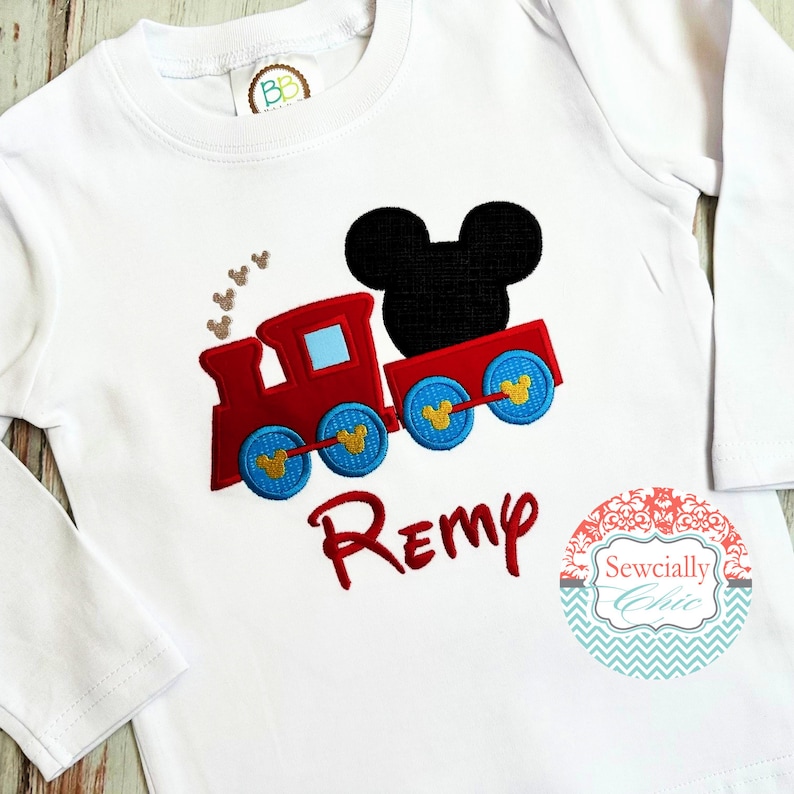 May include: White long-sleeved shirt with an embroidered red train with blue wheels and a black Mickey Mouse head. The train has gold Mickey Mouse ears on the wheels. The shirt has the name "Remy" embroidered in red below the train. The shirt also has a small circular label with the text "Sewcially Chic" in a decorative font.