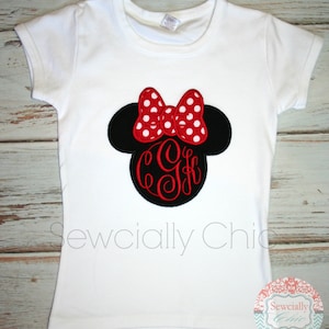 May include: White t-shirt featuring an embroidered Minnie Mouse head in black and red. The bow is red with white polka dots and there is a red monogram in the centre.