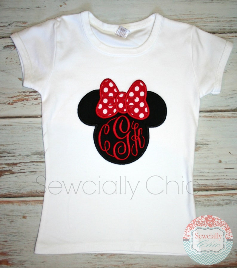 May include: White t-shirt featuring an embroidered Minnie Mouse head in black and red. The bow is red with white polka dots and there is a red monogram in the centre.